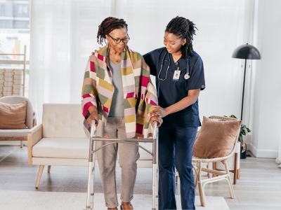 A caretaker helping an elderly woman walk. The elderly woman is using a walker.